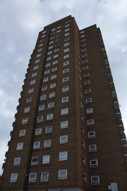 A close-up view of a multi-storey residential building with numerous balconies made of concrete and metal railings, some with plants, laundry, and outdoor furniture. The building’s exterior features